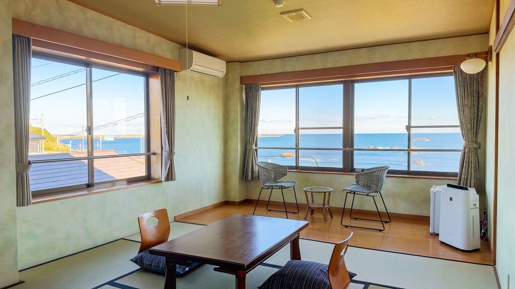 Habitació amb taula de fusta, dues cadires, taula rodona petita, nevera i finestres grans amb vista al mar.