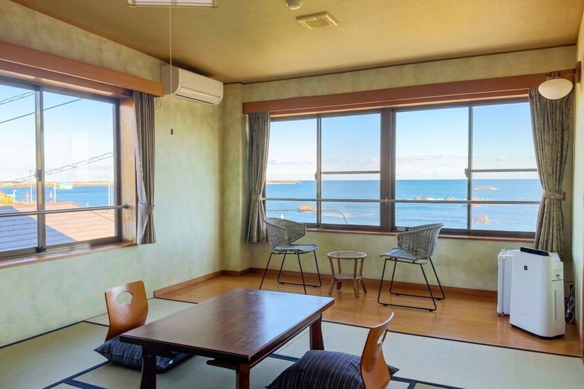 A room with a wooden table, two chairs, a small round table, a refrigerator, and large windows with a view of the sea.