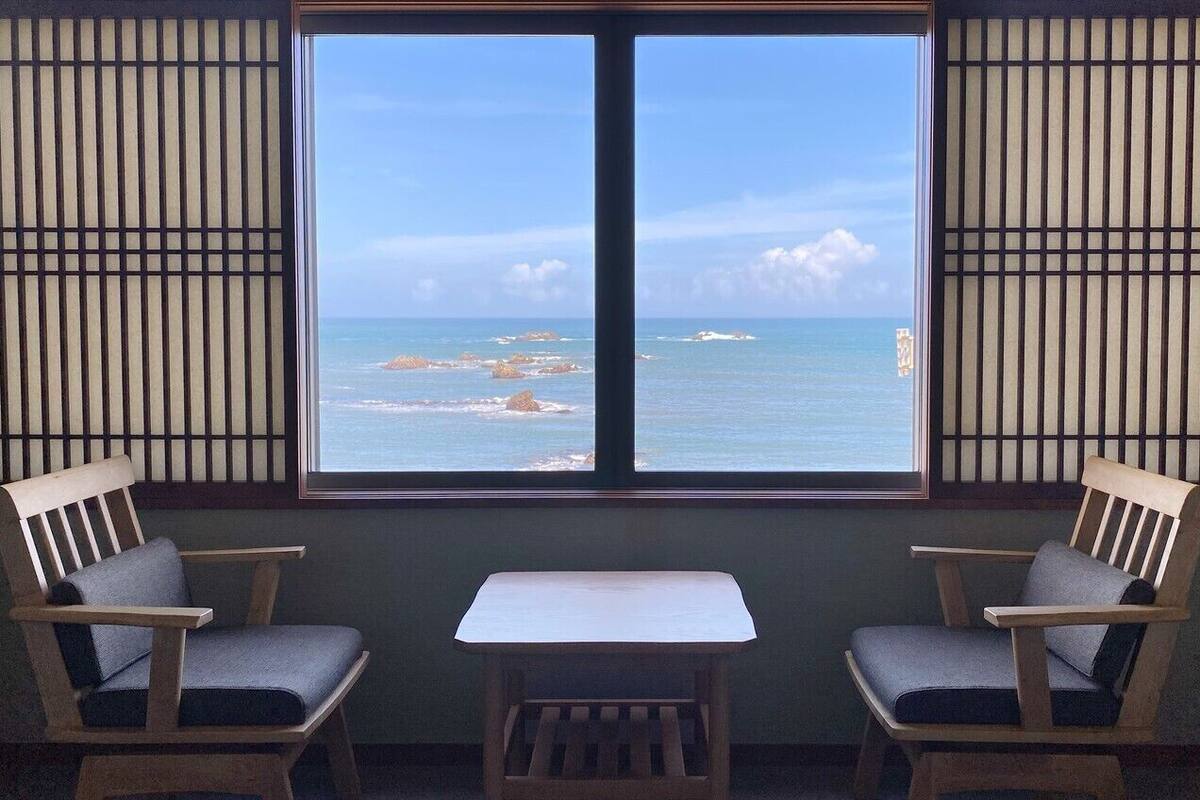A room with a view of the ocean through a large window, two wooden chairs, and a small table.