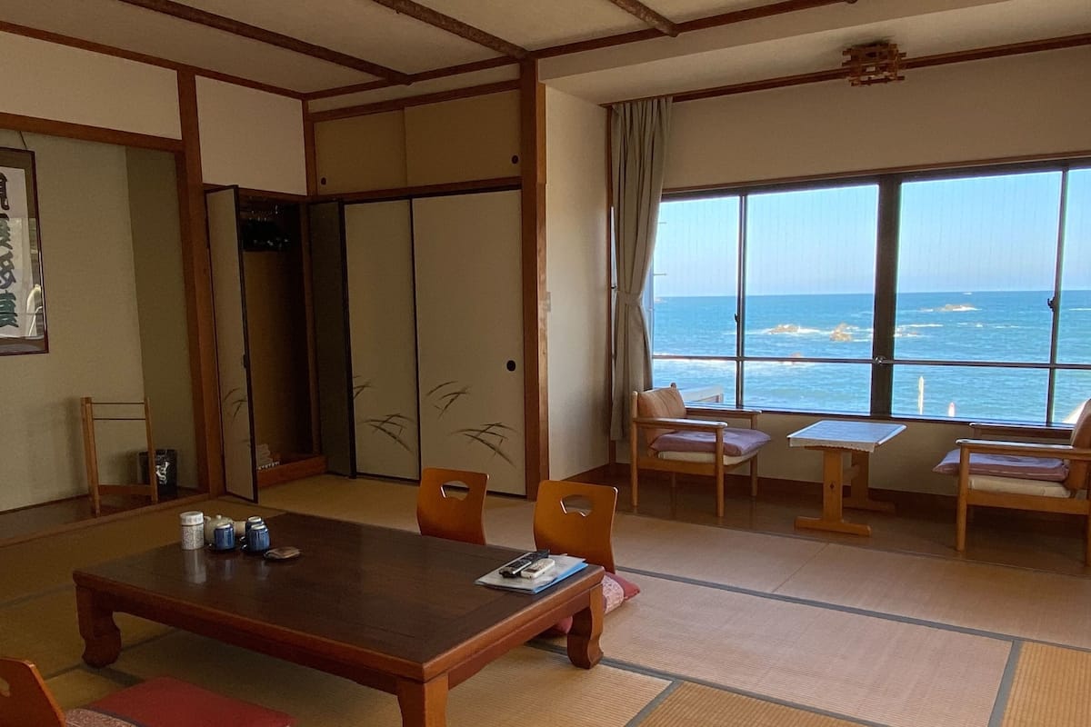 A traditional Japanese room with tatami flooring, a low table, chairs, a television, and a view of the sea through large windows.