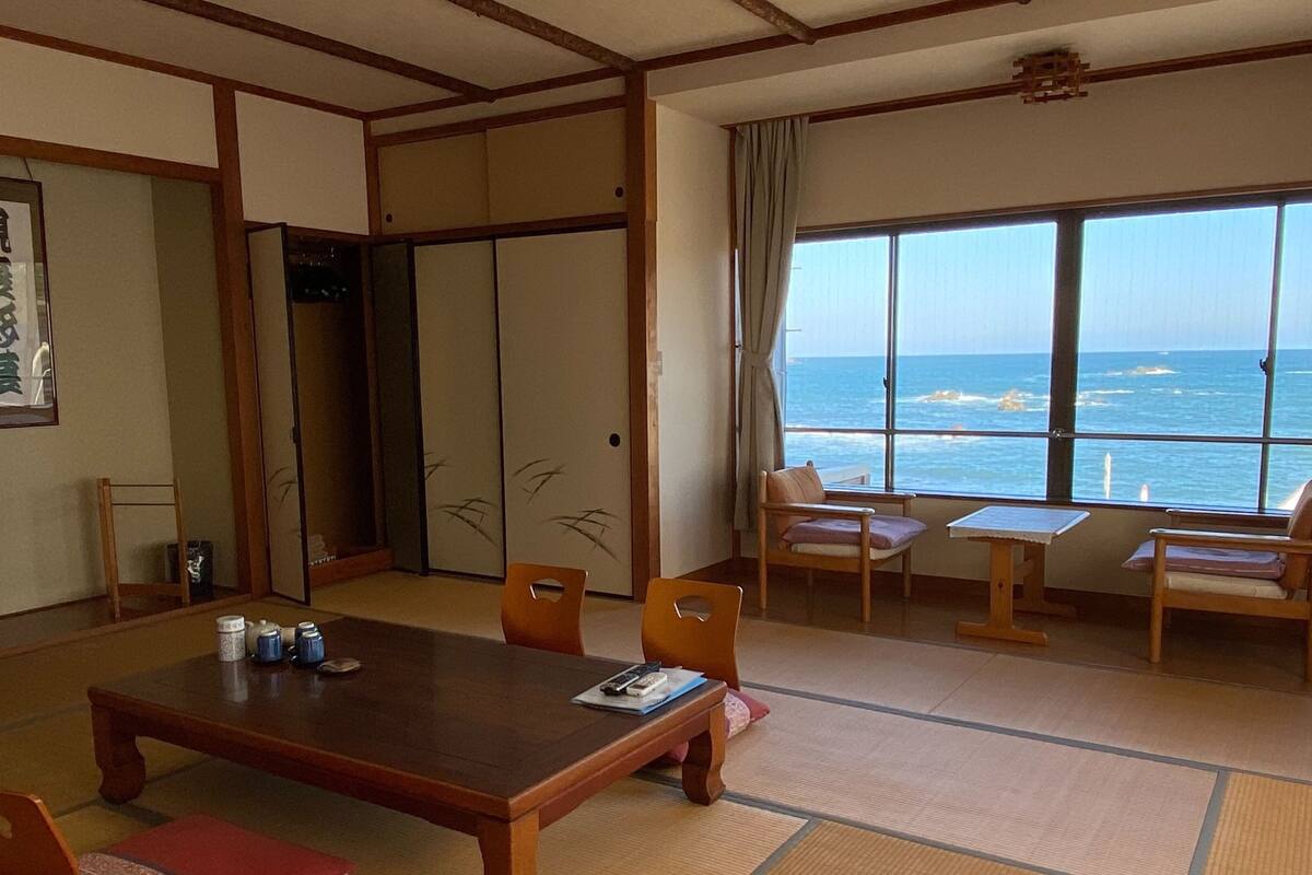 Une pièce de style japonais traditionnel, avec un sol recouvert de tatamis, une table basse, des chaises, un téléviseur et une vue sur la mer grâce à de grandes fenêtres.