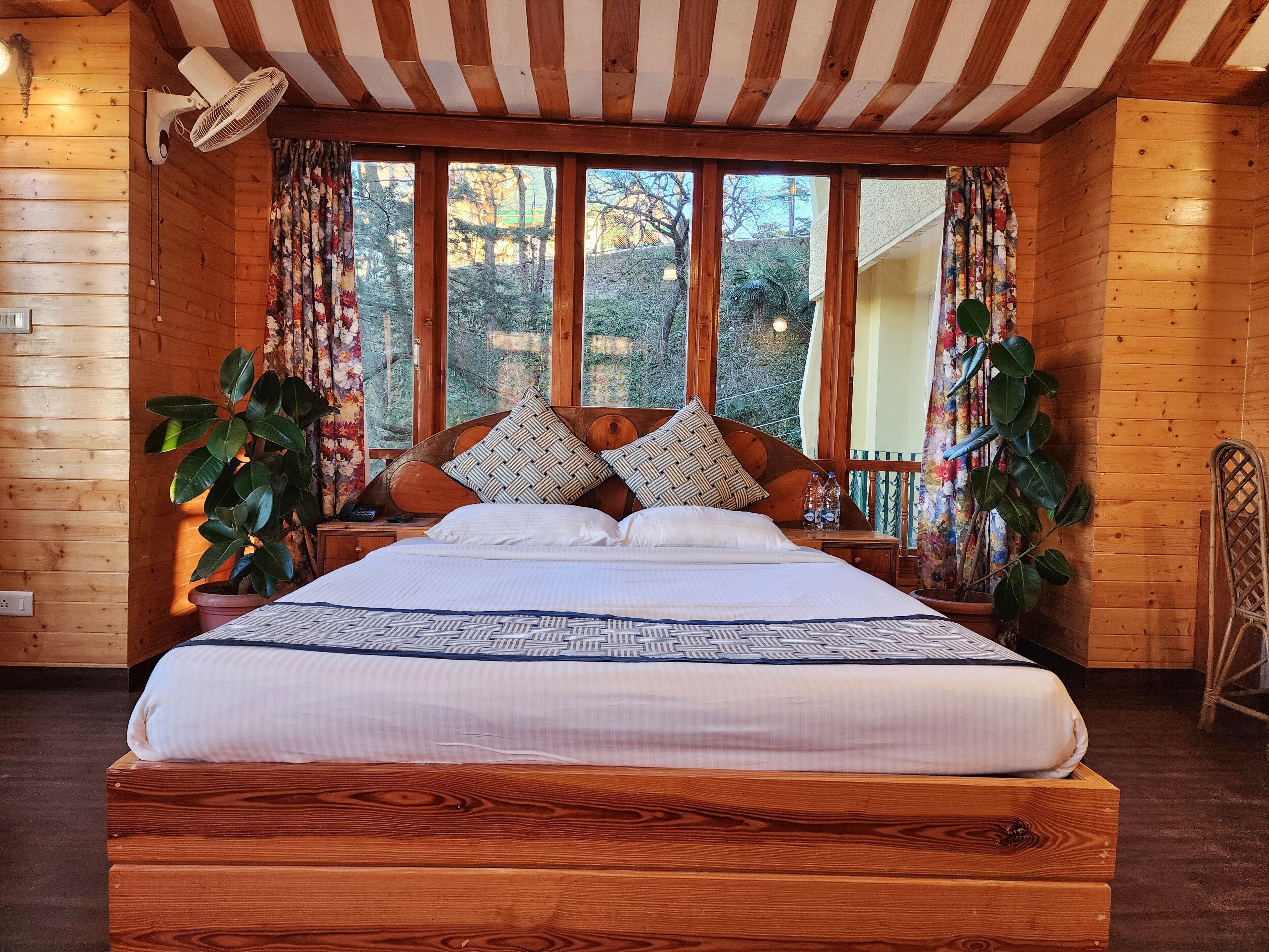A wooden cabin bedroom with a large bed, two potted plants, and a view of trees through large windows.