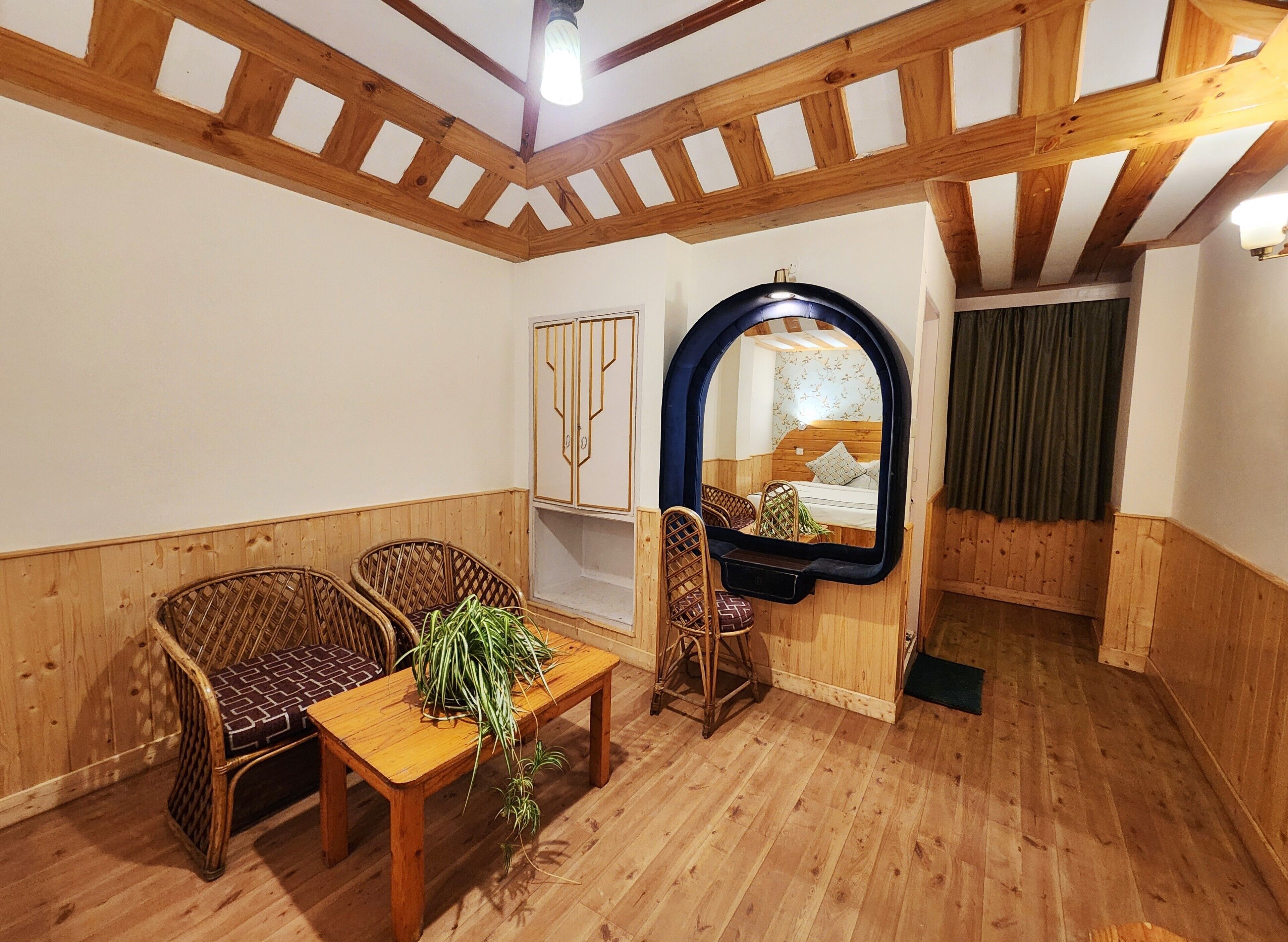 Une pièce au plancher de bois, avec deux fauteuils en osier, une petite table sur laquelle est posée une plante, et un miroir qui reflète un lit.