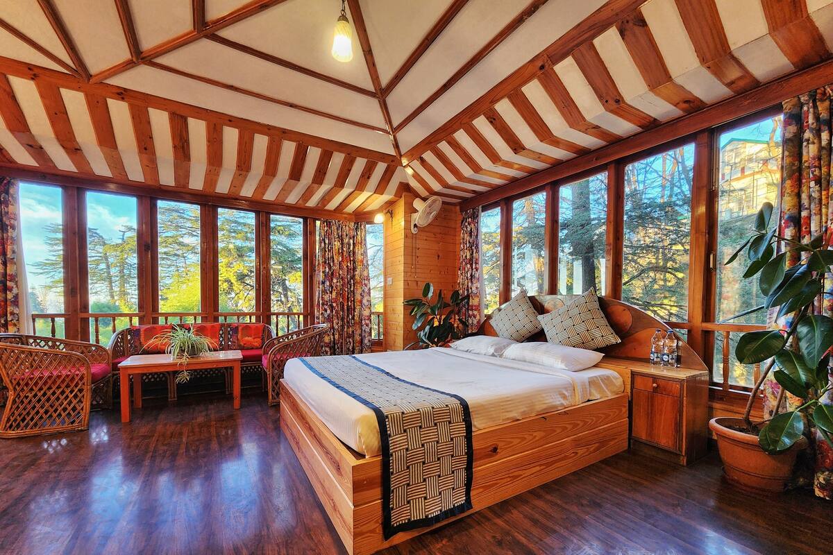 Quarto espaçoso com cama grande, soalho de madeira e amplas janelas com vista para árvores e edifícios.