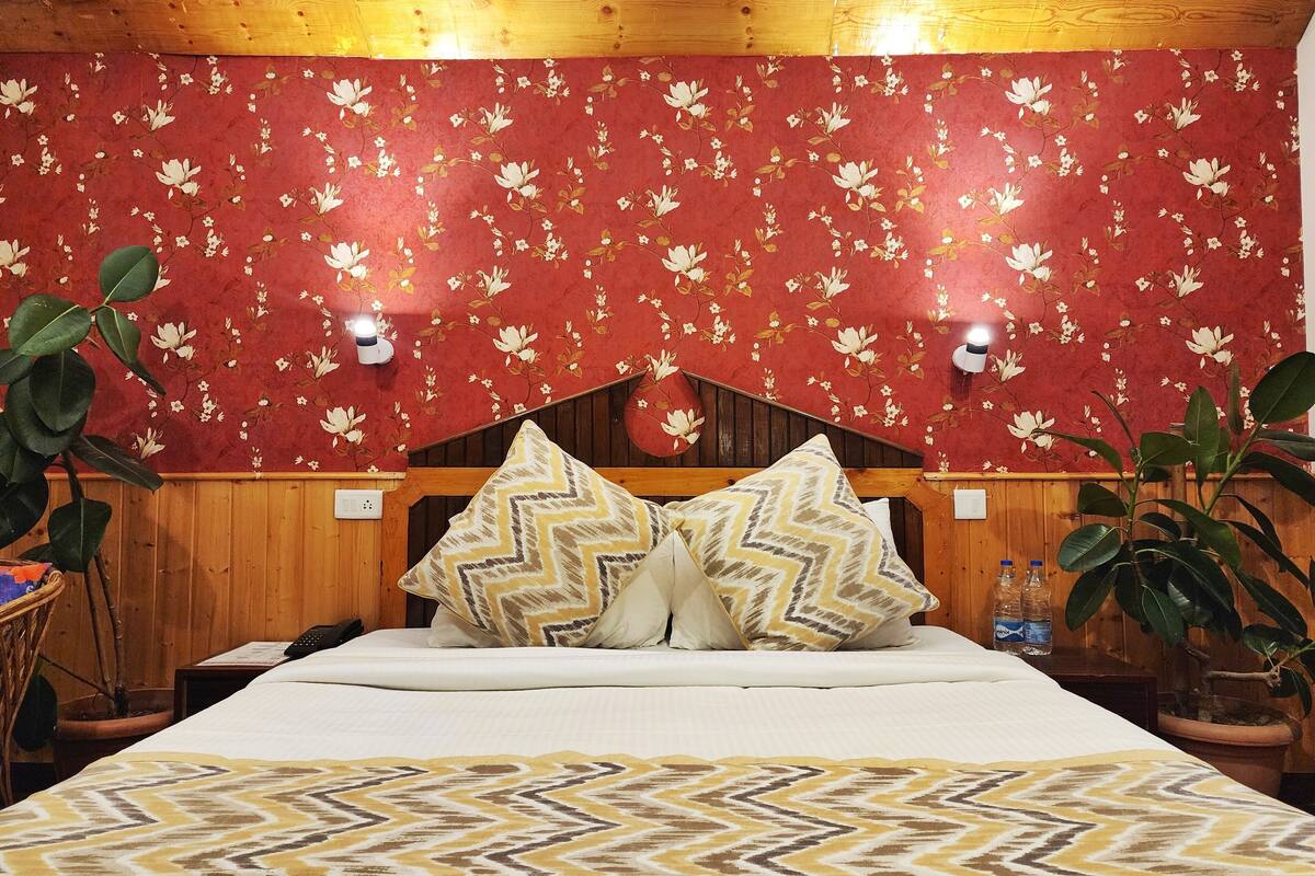 Quarto com cabeceira de madeira, papel de parede floral, duas mesas de cabeceira, cama com almofadas estampadas e plantas em vasos.