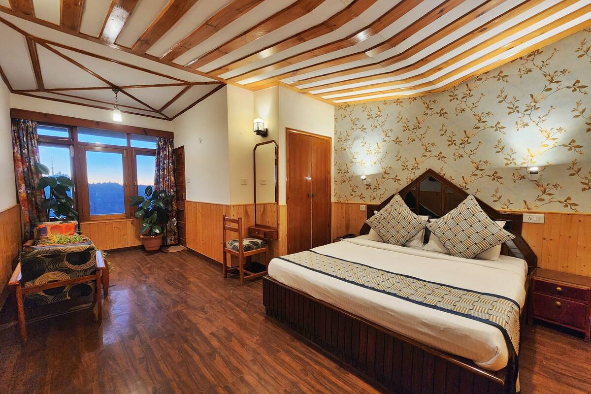 Um quarto aconchegante com uma cama grande, vigas de madeira no teto, uma janela com cortinas, uma cadeira e uma mesa de cabeceira.