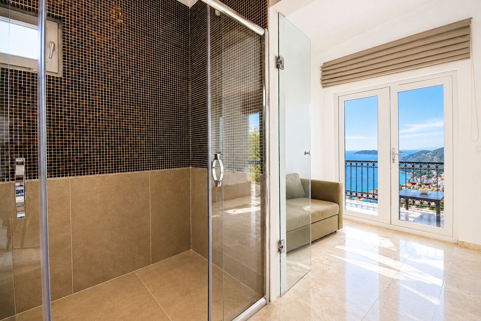A modern bathroom with a glass shower enclosure, a beige tiled wall, and a glass door leading to a balcony with a view of the sea.