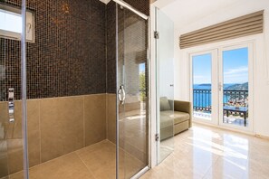 Un baño moderno con cabina de ducha de vidrio, pared con azulejos color beige y puerta de vidrio que da a un balcón con vista al mar.