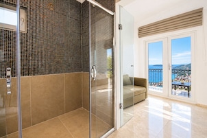 Un baño moderno con cabina de ducha de vidrio, pared con azulejos color beige y puerta de vidrio que da a un balcón con vista al mar.