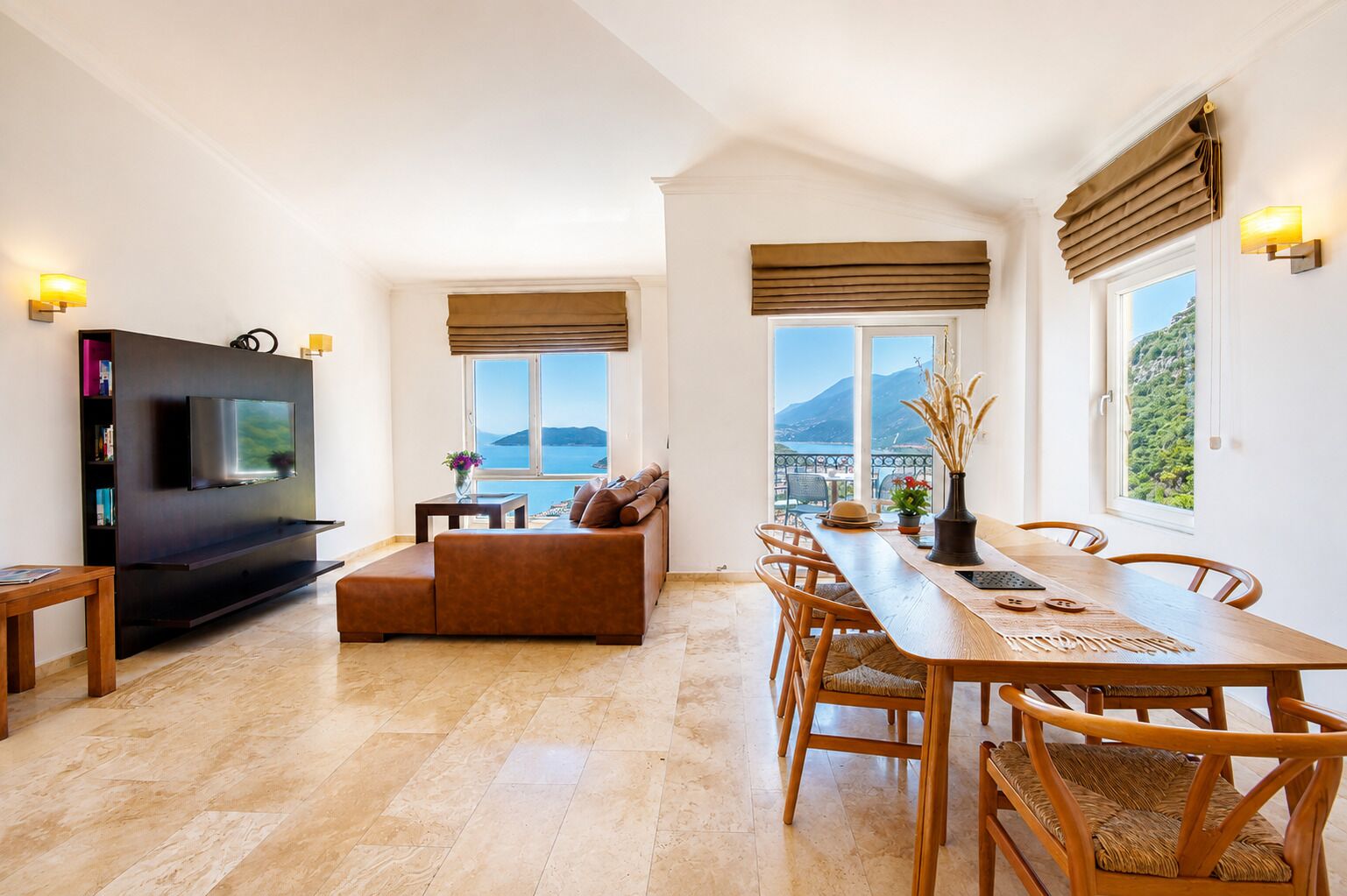 A modern living room with a flat-screen TV, a sofa, a dining table with chairs, and large windows with views of the sea and mountains.