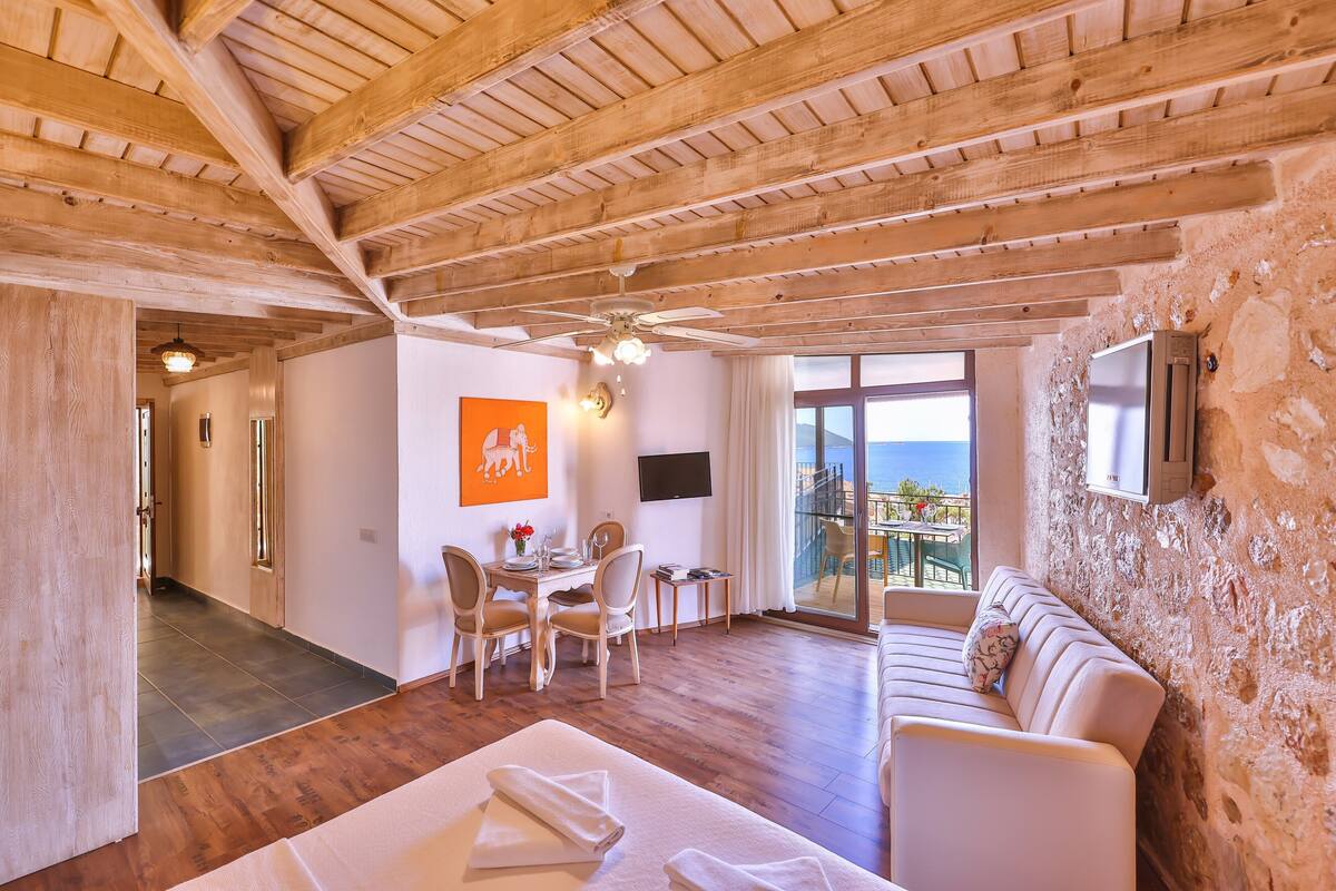 A room with wooden beams, a sofa, a dining area, and a balcony with a view.