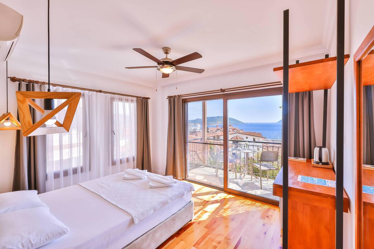 A hotel room with a bed, a ceiling fan, a wooden desk, and a balcony with a view of the city.
