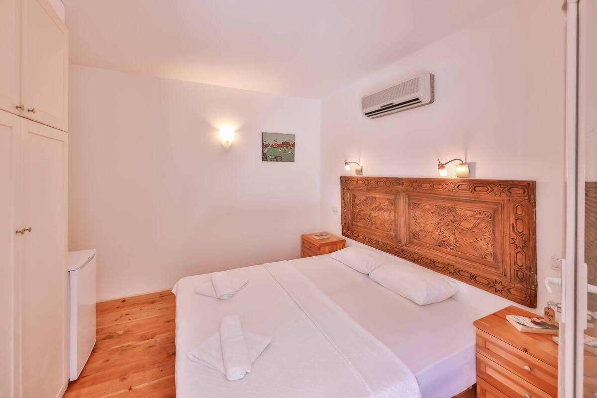 A hotel room with a large bed, a wooden headboard, a wall-mounted air conditioner, and a small bedside table with a lamp.