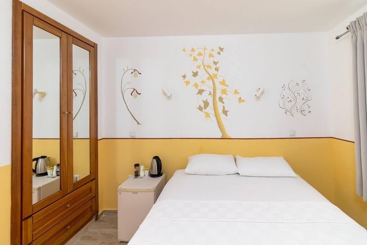 A hotel room with a bed, a wardrobe, a small bedside table with a kettle, and a wall with a tree and bird decals.