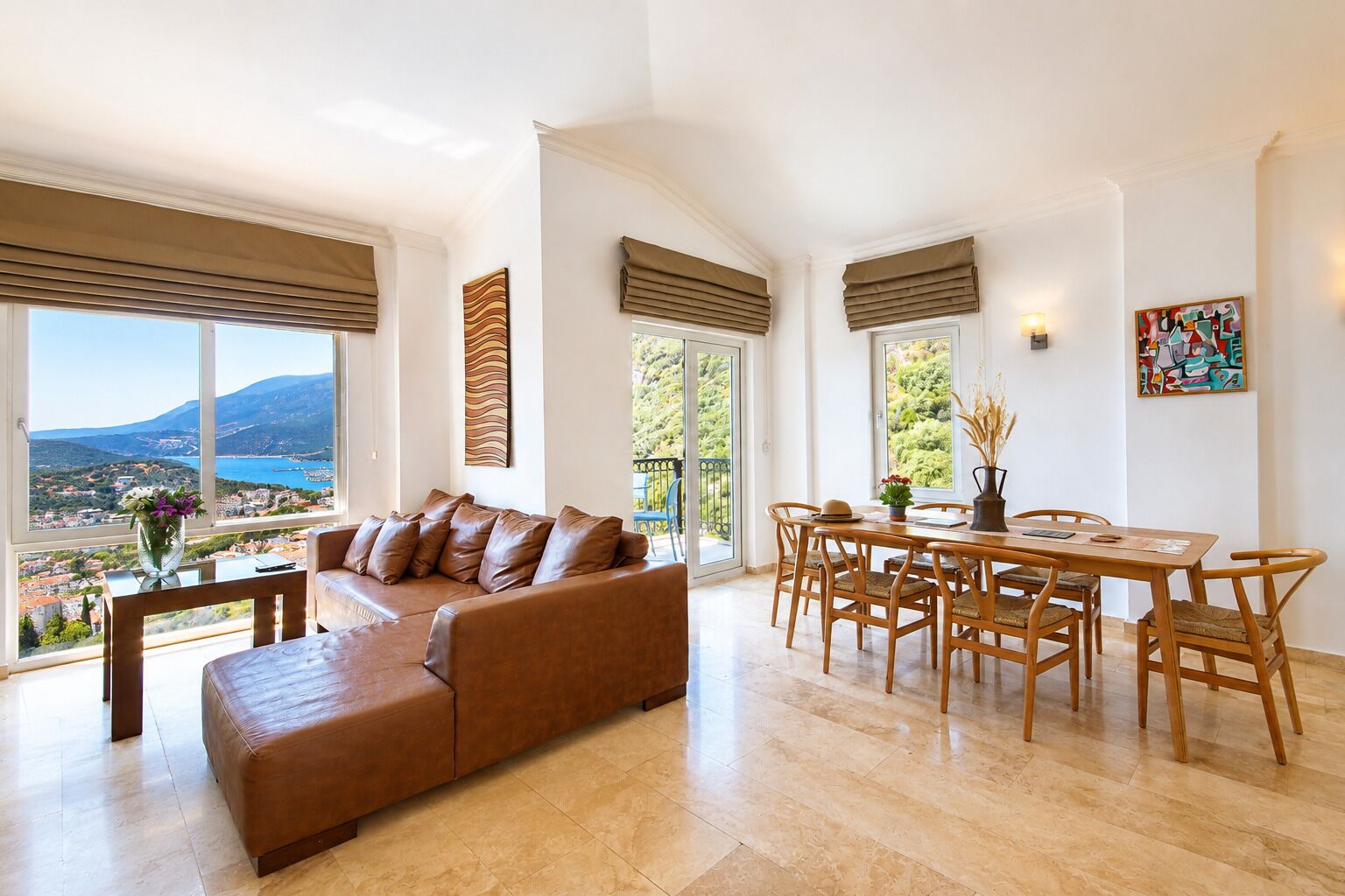 A spacious living room with a large window offering a scenic view, a brown leather sofa, a dining table with chairs, and a painting on the wall.