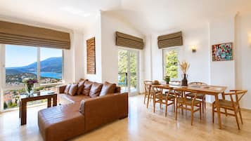 A spacious living room with a large window offering a scenic view, a brown leather sofa, a dining table with chairs, and a painting on the wall.
