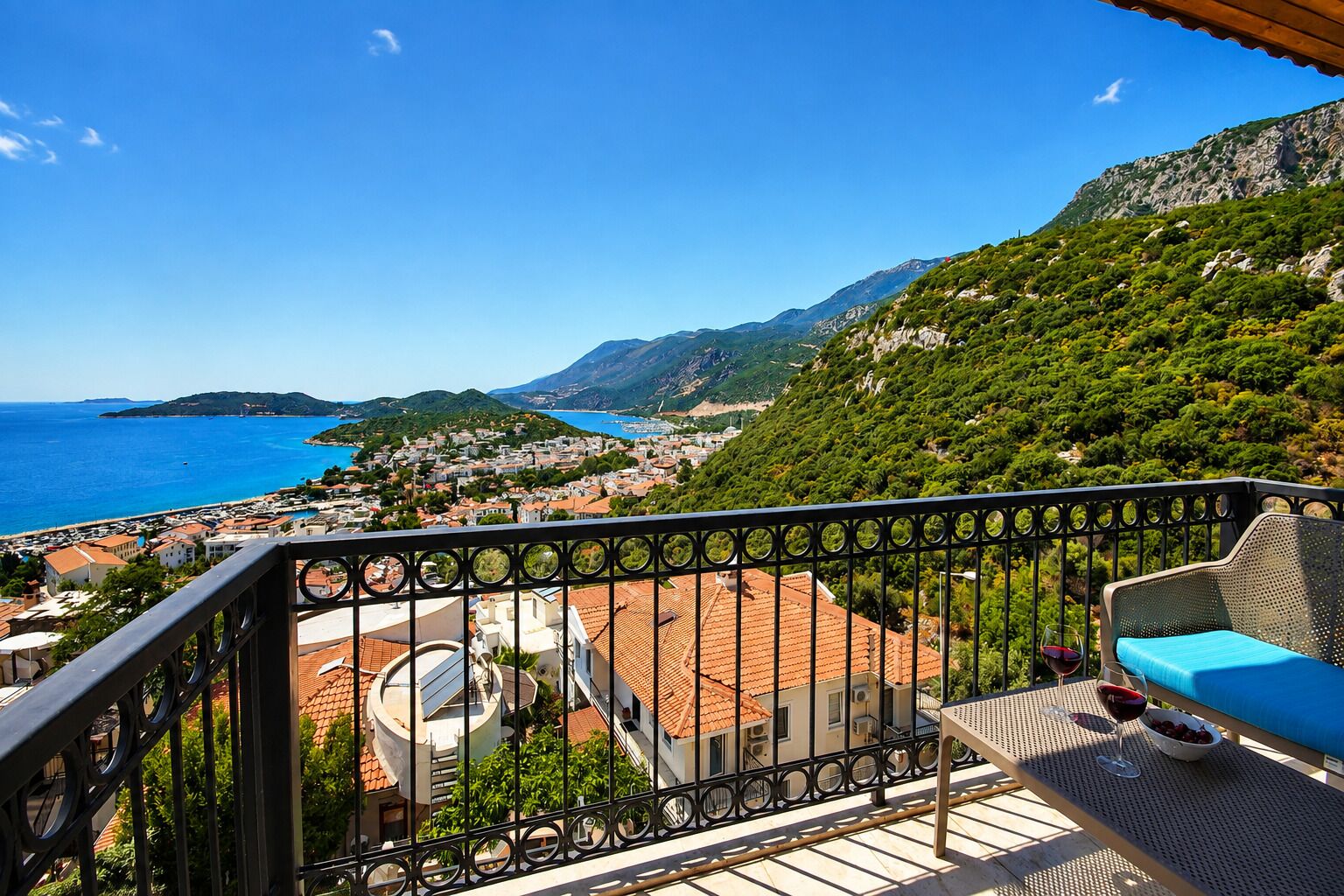 A balcony with a view of a coastal town, a table with a glass of red wine, and a blue cushion.