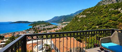 A balcony with a view of a coastal town, a table with a glass of red wine, and a blue cushion.