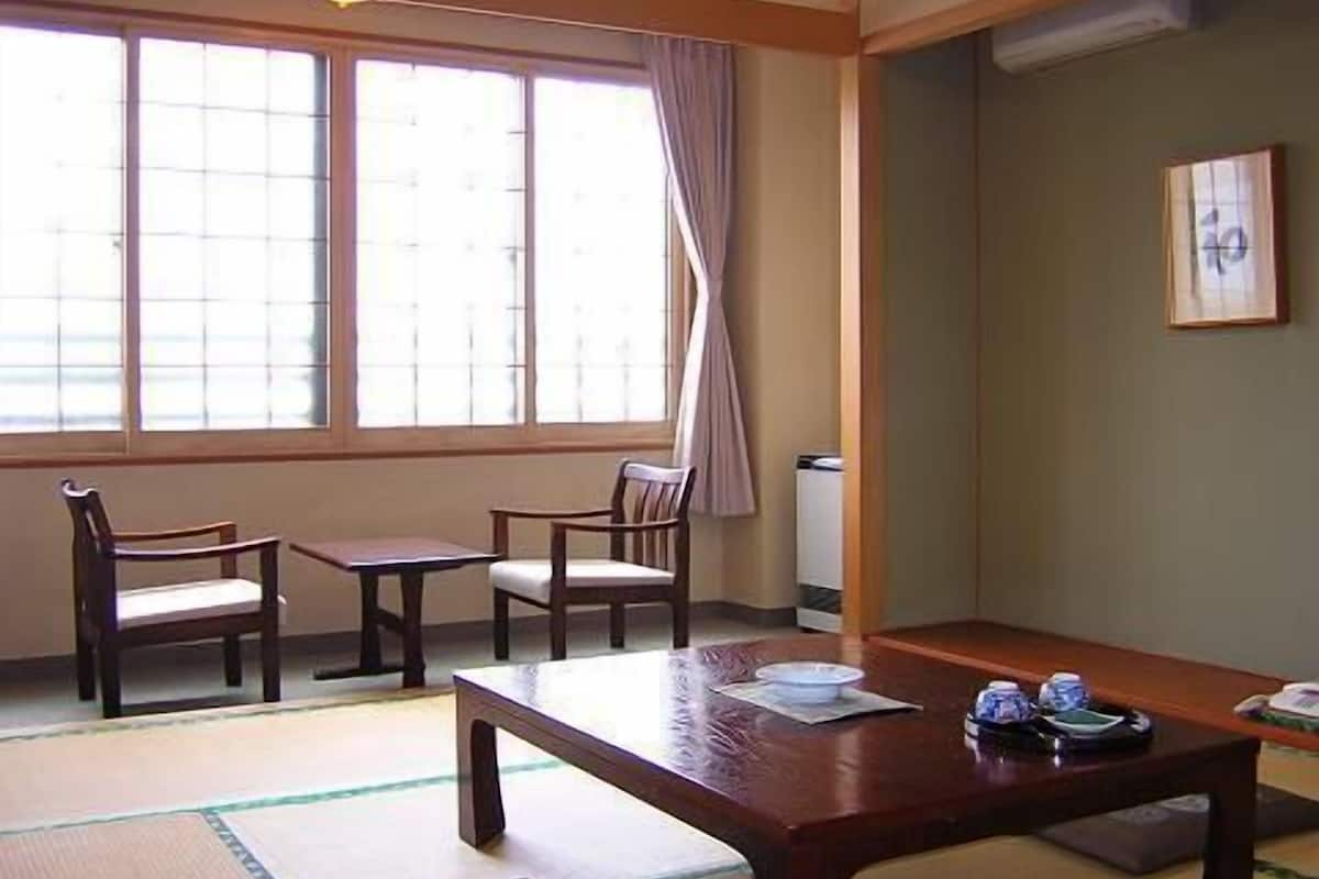 A traditional Japanese room with tatami flooring, a low wooden table, two chairs, and a large window with curtains.
