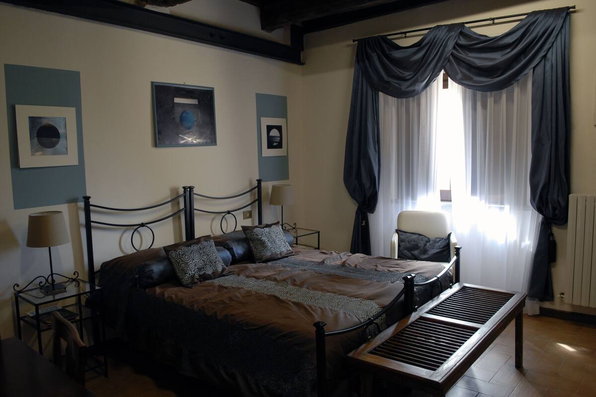 Une chambre à coucher avec un grand lit, une table de chevet, une chaise et une fenêtre avec des rideaux.