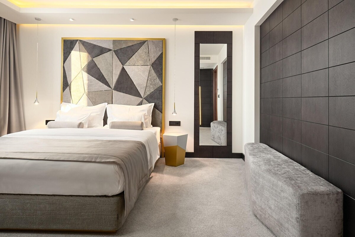 A modern bedroom with a large bed, a geometric wall art piece, a bench, and a mirror.