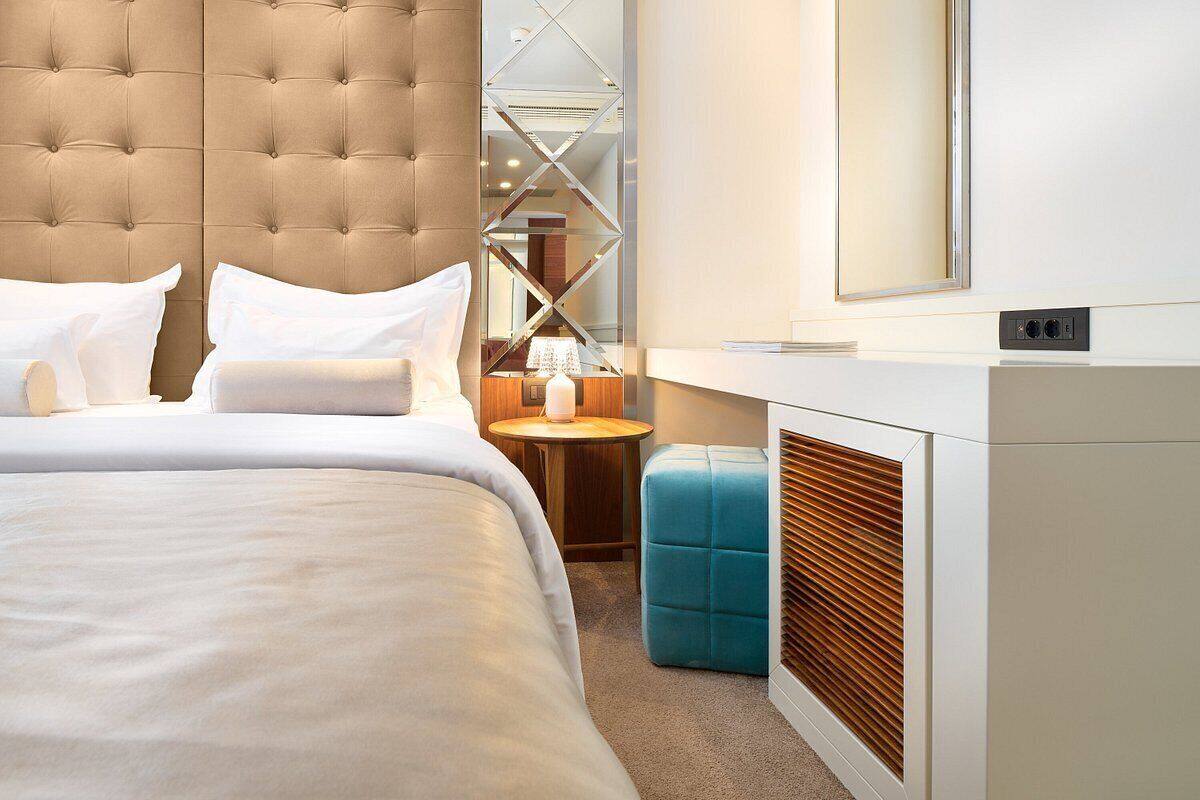 A modern hotel room with a tufted headboard, a large bed, a small round table, a blue ottoman, and a built-in heater.