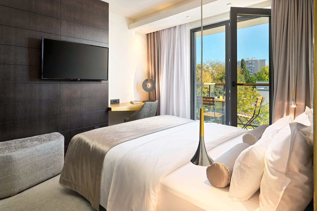 A modern hotel room with a large bed, a flat-screen TV mounted on the wall, a balcony with a view of the city, and a desk with a lamp.