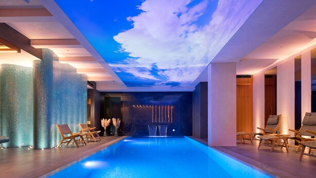 Ein modernes Hallenbad mit einer Decke, die Himmel und Wolken darstellt, umgeben von Liegestühlen und einer Holzverkleidung.