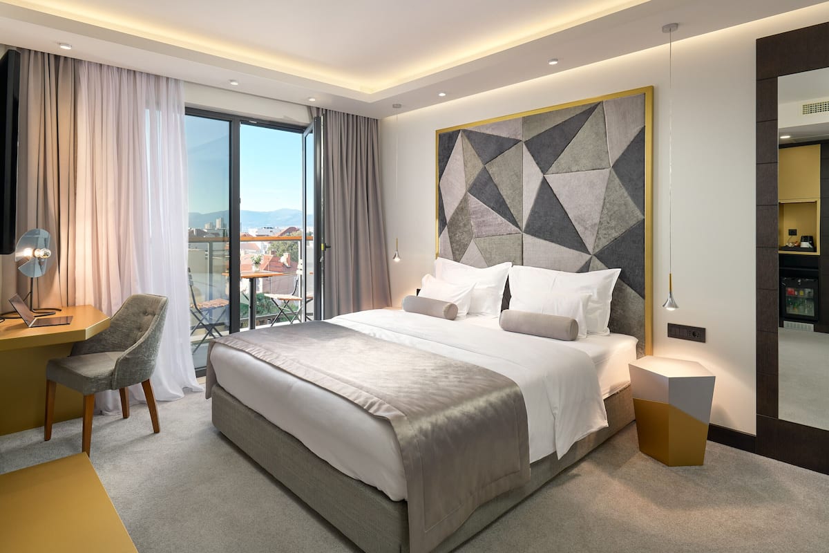 A modern hotel room with a large bed, a desk with a chair, a bedside table, and a view of the city through the window.