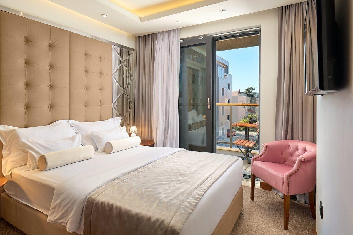 A hotel room with a large bed, a pink chair, a TV, and a balcony with a view of buildings.