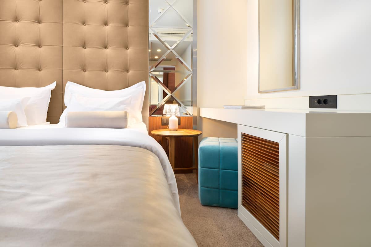 A modern hotel room with a tufted headboard, a large bed with white linens, a small round table with a lamp, a blue ottoman, and a built-in heater.