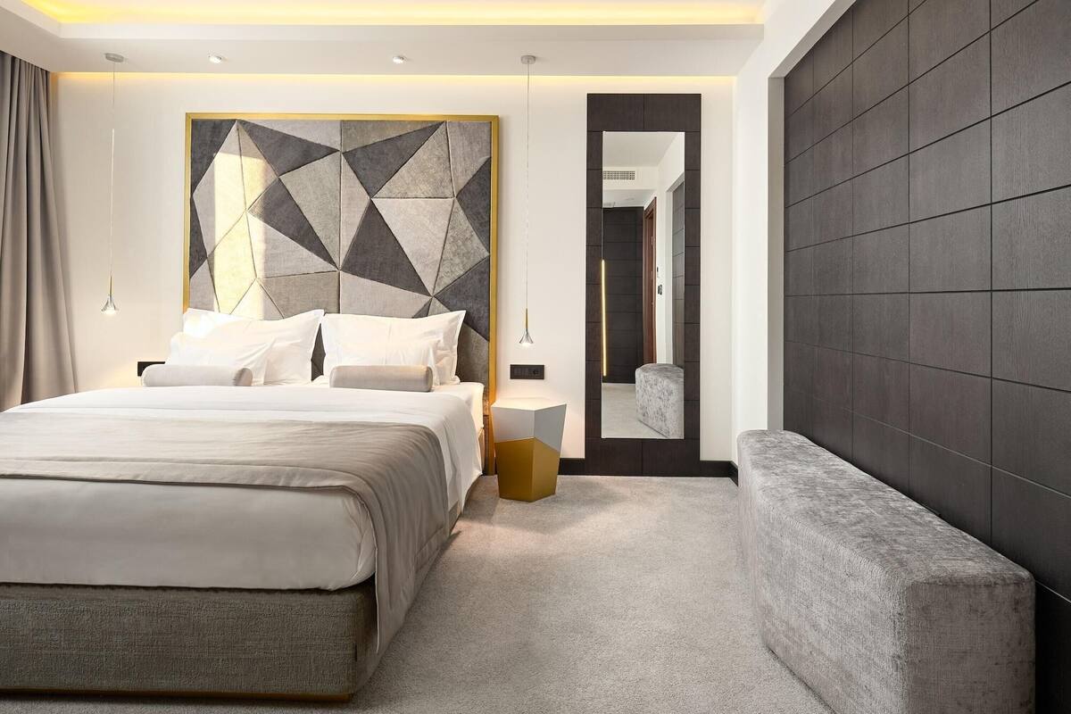 A modern bedroom with a large bed, a geometric wall art piece, a bench, and a mirror.