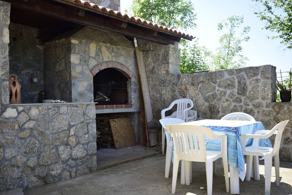 Forno in pietra all'aperto con arco in mattoni, circondato da sedie di plastica bianca e un tavolo con tovaglia blu.