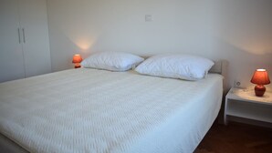 Una cama bien hecha con ropa blanca y dos almohadas, flanqueada por dos mesitas de noche con lámparas, en una habitación sencilla con paredes blancas y suelo de madera.