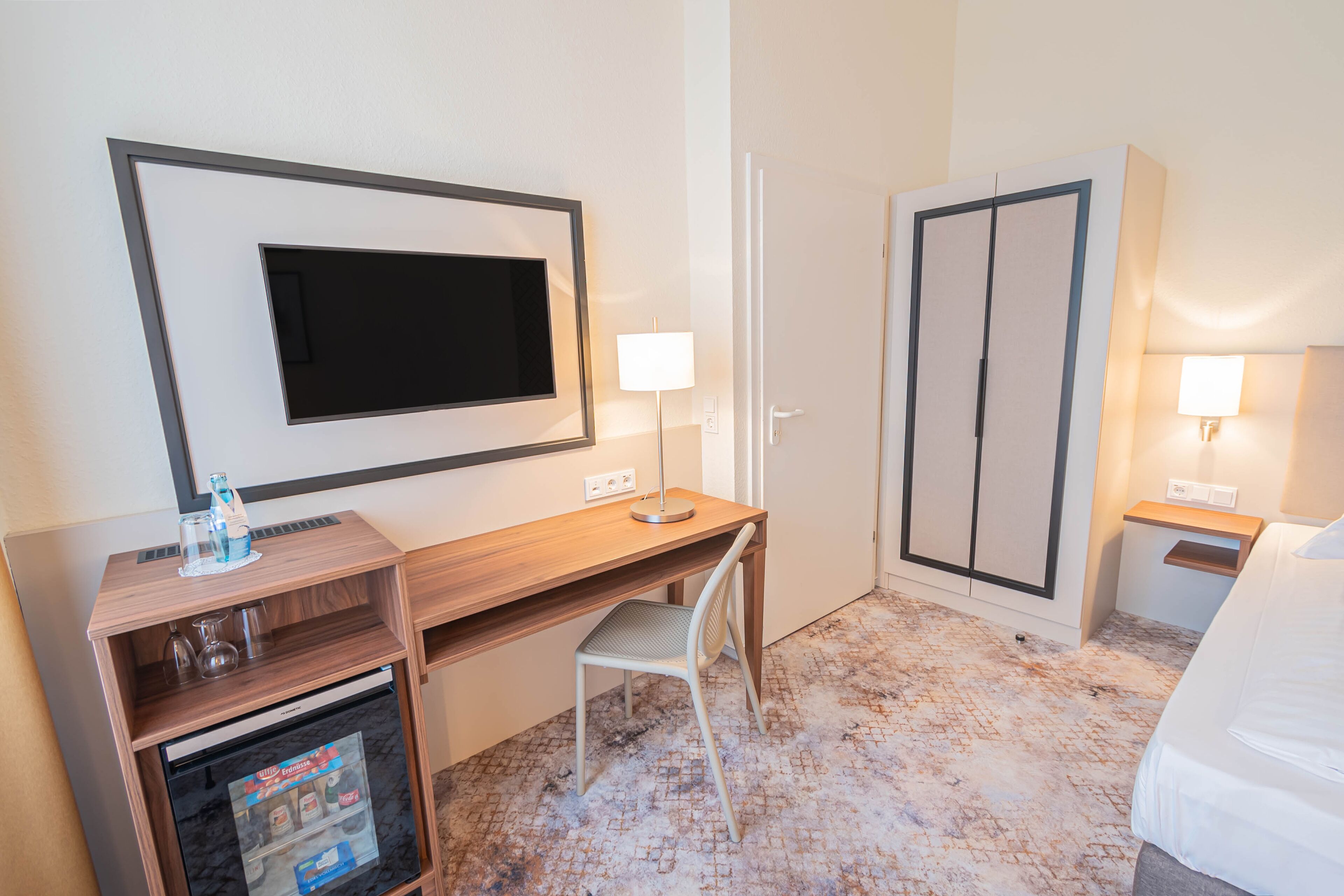 A hotel room with a wooden desk, a chair, a television mounted on the wall, a wardrobe, and a bed.