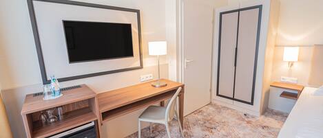 A hotel room with a wooden desk, a chair, a television mounted on the wall, a wardrobe, and a bed.