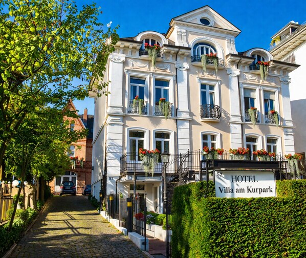 Ein Hotelgebäude mit mehreren Balkonen, einer Kopfsteinpflasterstraße und einer Hecke davor.