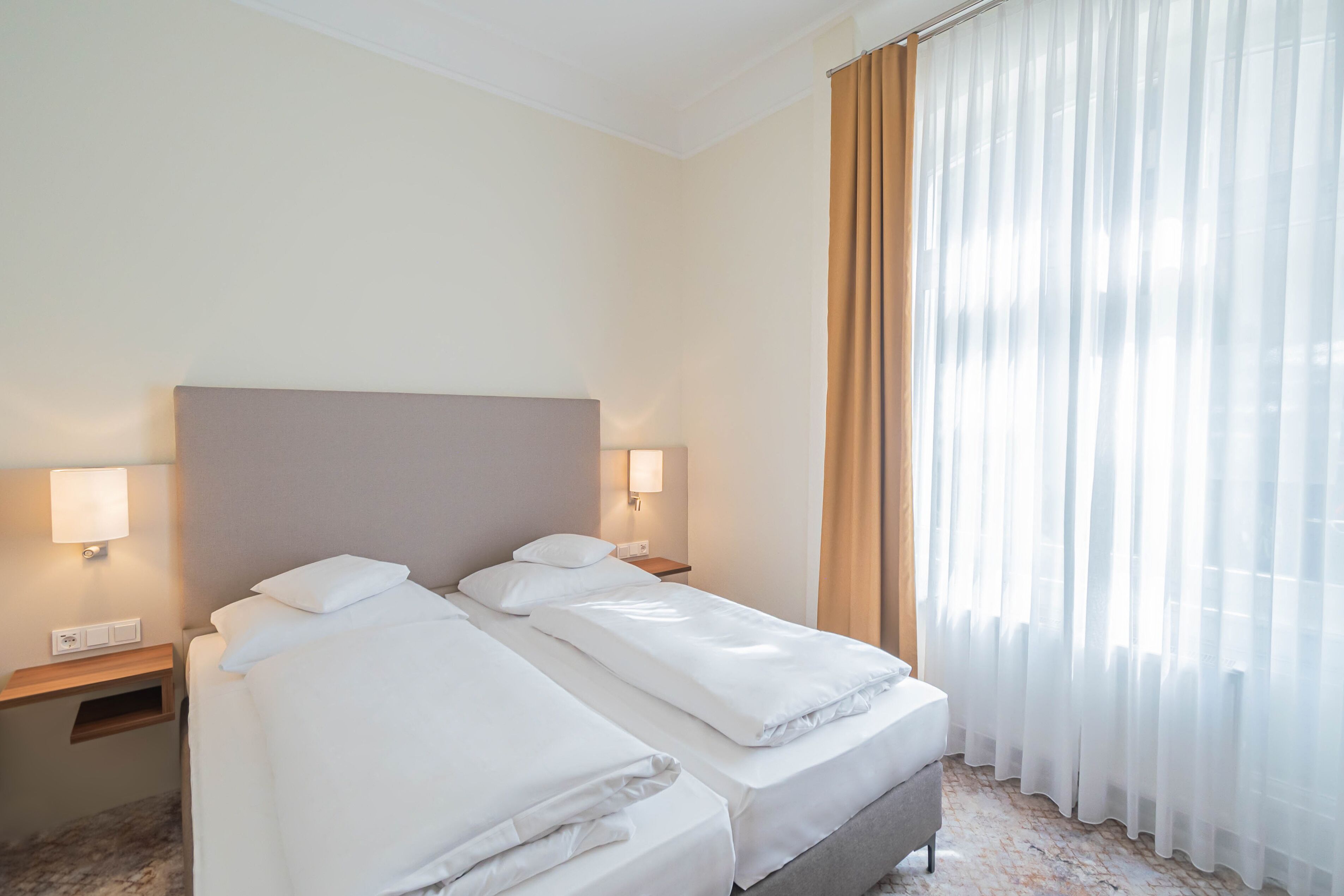 A hotel room with two beds, a headboard, bedside lamps, and a window with sheer curtains.