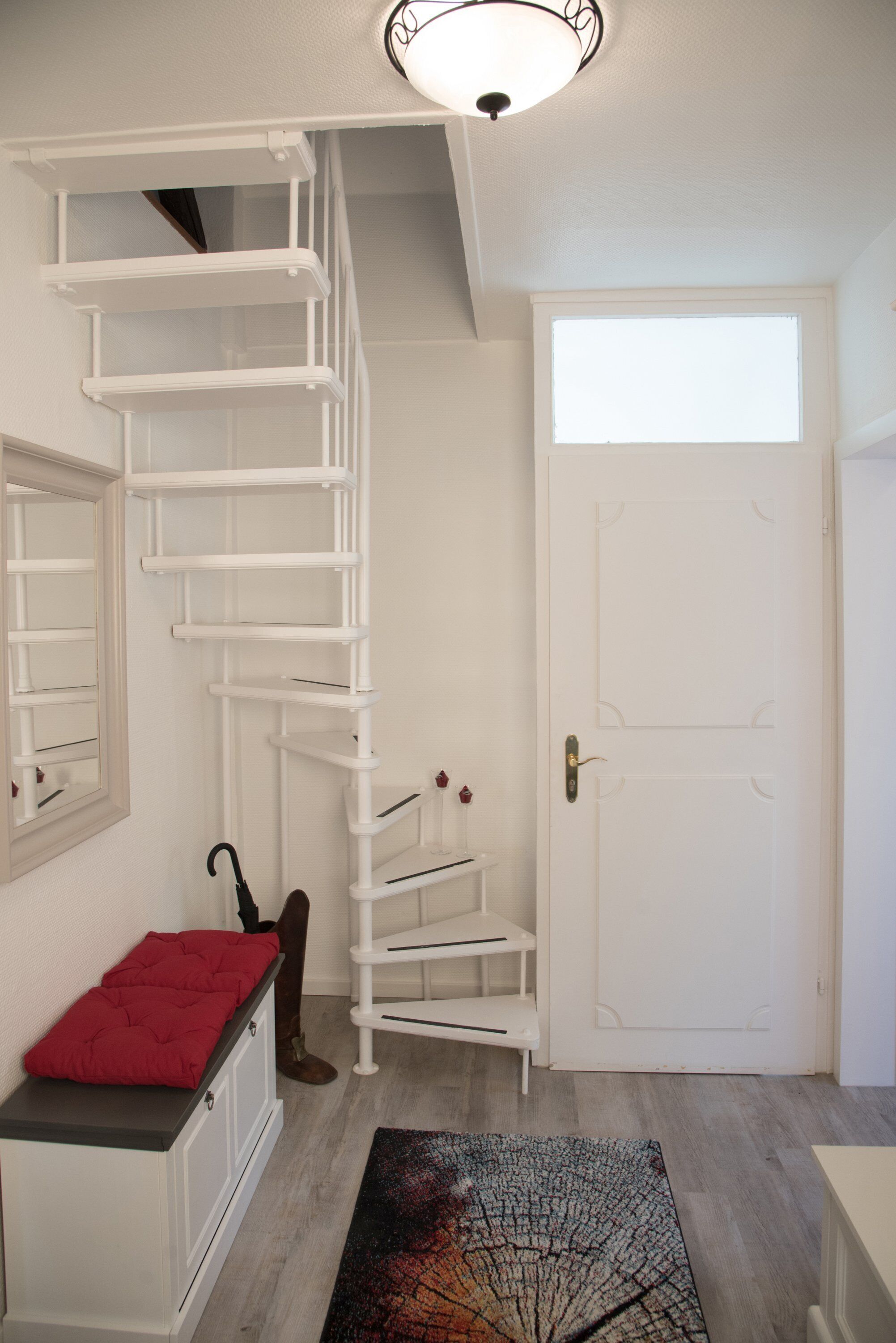 Un escalier menant à un deuxième étage, une porte blanche et un coussin rouge posé sur un meuble blanc.