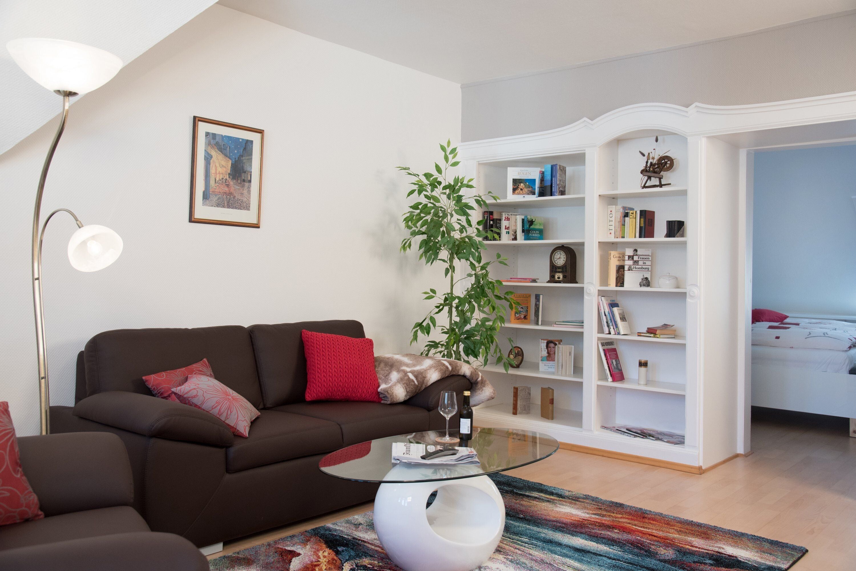 A modern living room with a brown sofa, a glass coffee table, a bookshelf, and a plant.
