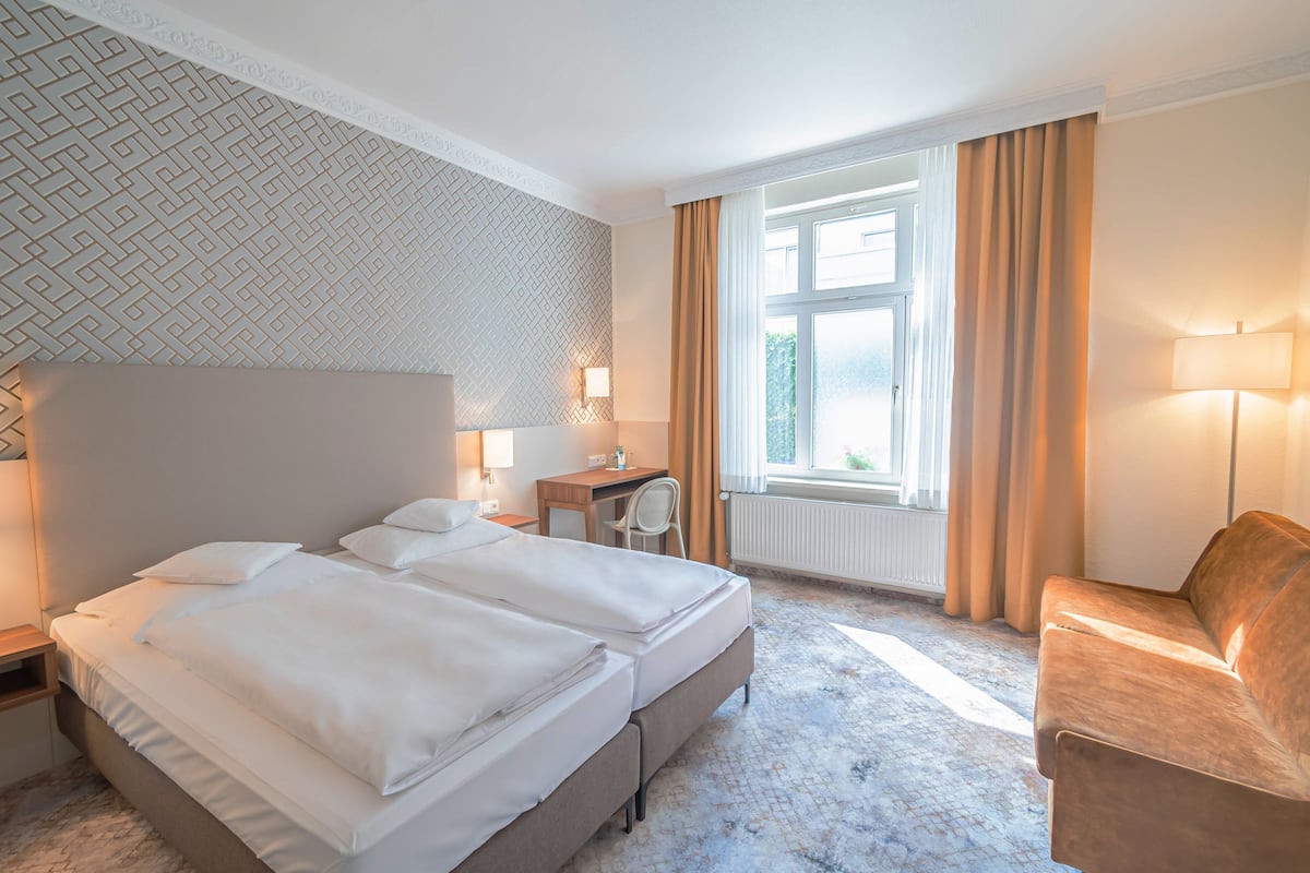 A hotel room with two beds, a desk, a chair, and a large window with curtains.