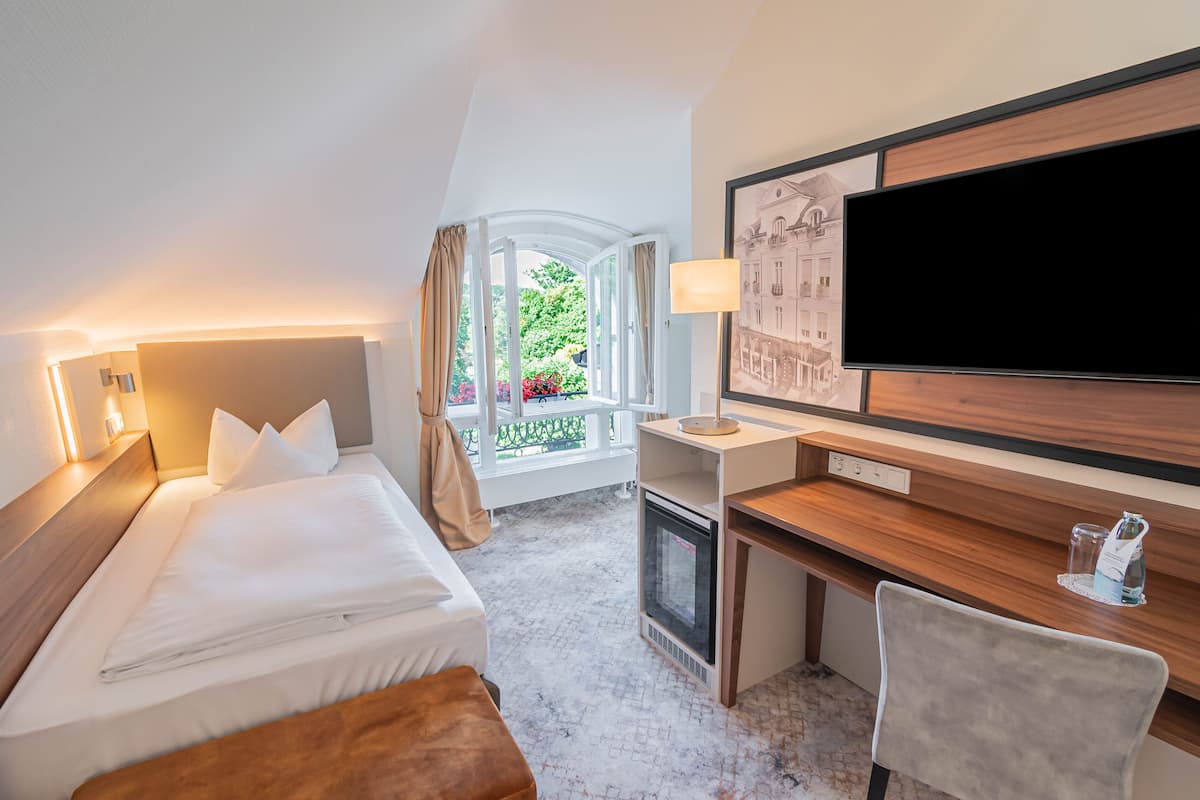 A hotel room with a bed, desk, TV, and a view of greenery outside.