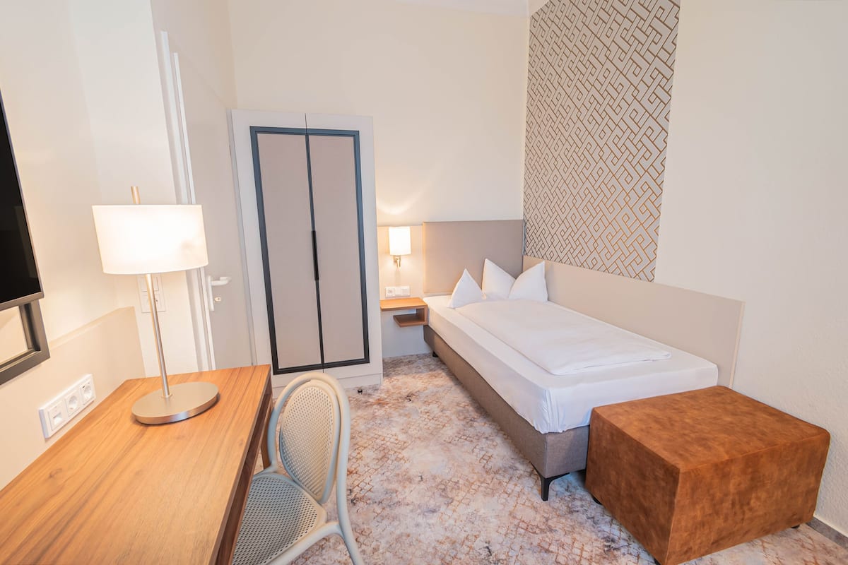 A hotel room with a bed, bedside table, lamp, chair, and a small wooden stool.