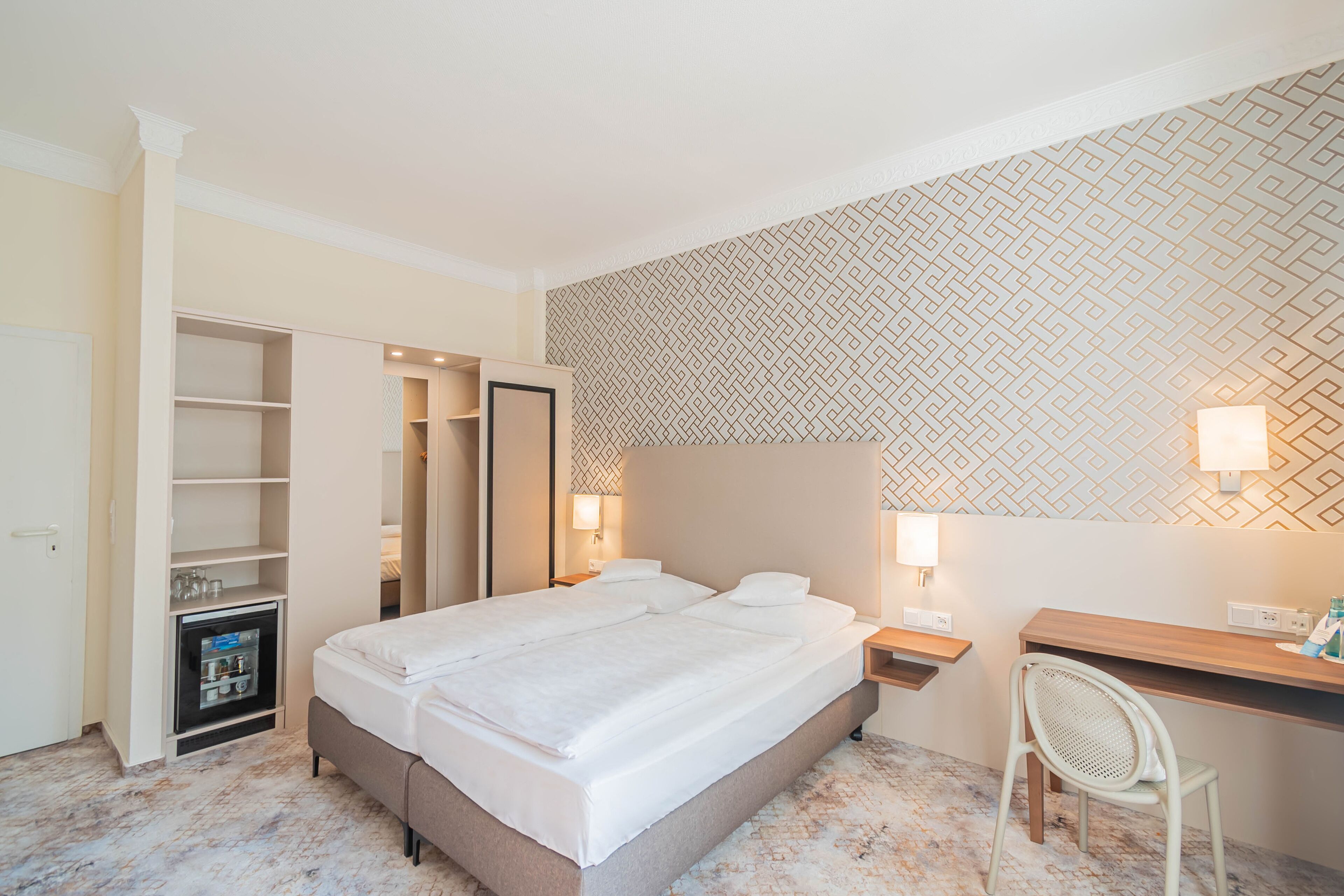 A hotel room with a large bed, a desk, a chair, a wardrobe, and a minibar.