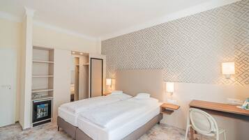 A hotel room with a large bed, a desk, a chair, a wardrobe, and a minibar.