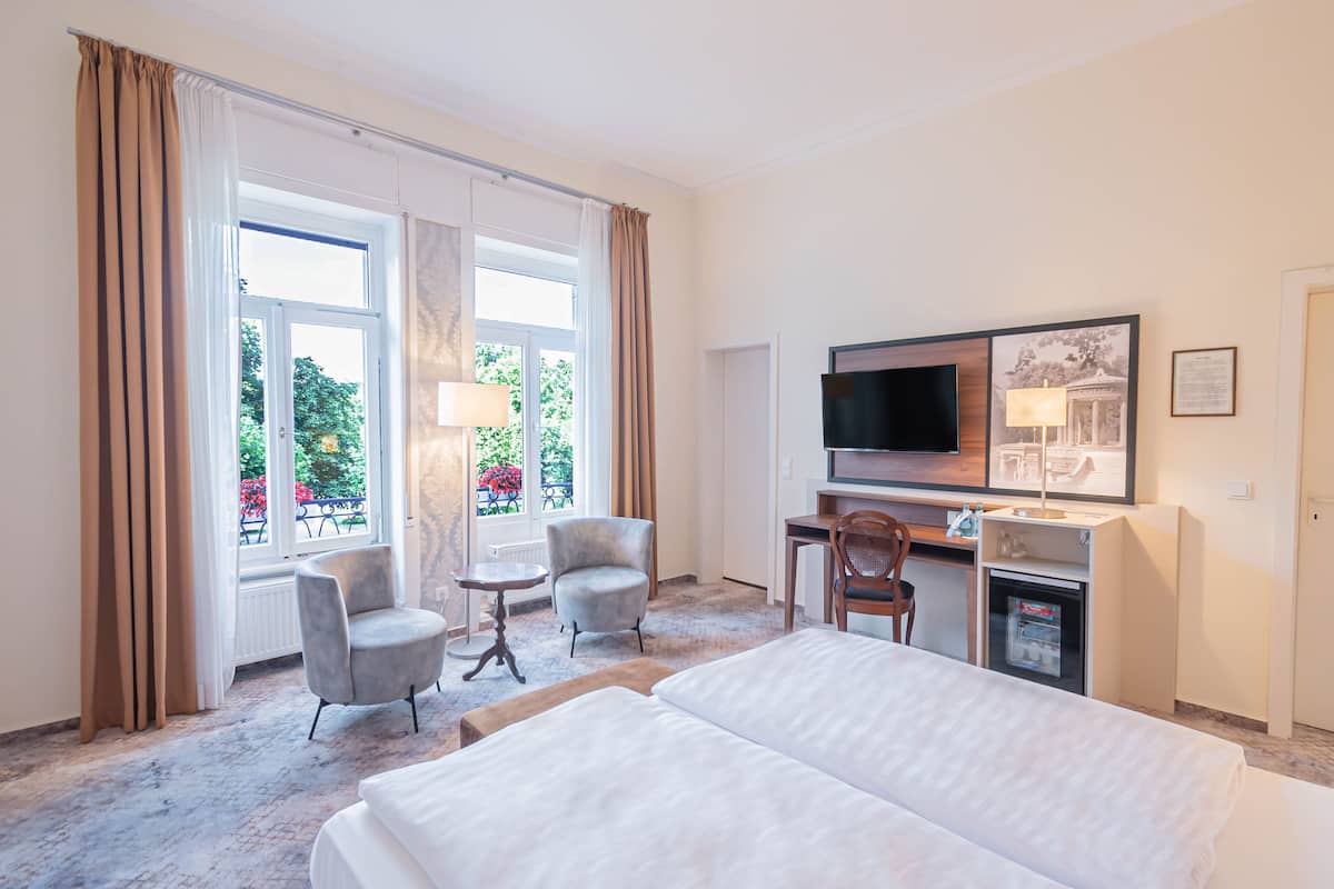A hotel room with a bed, two armchairs, a desk with a television, and a refrigerator.