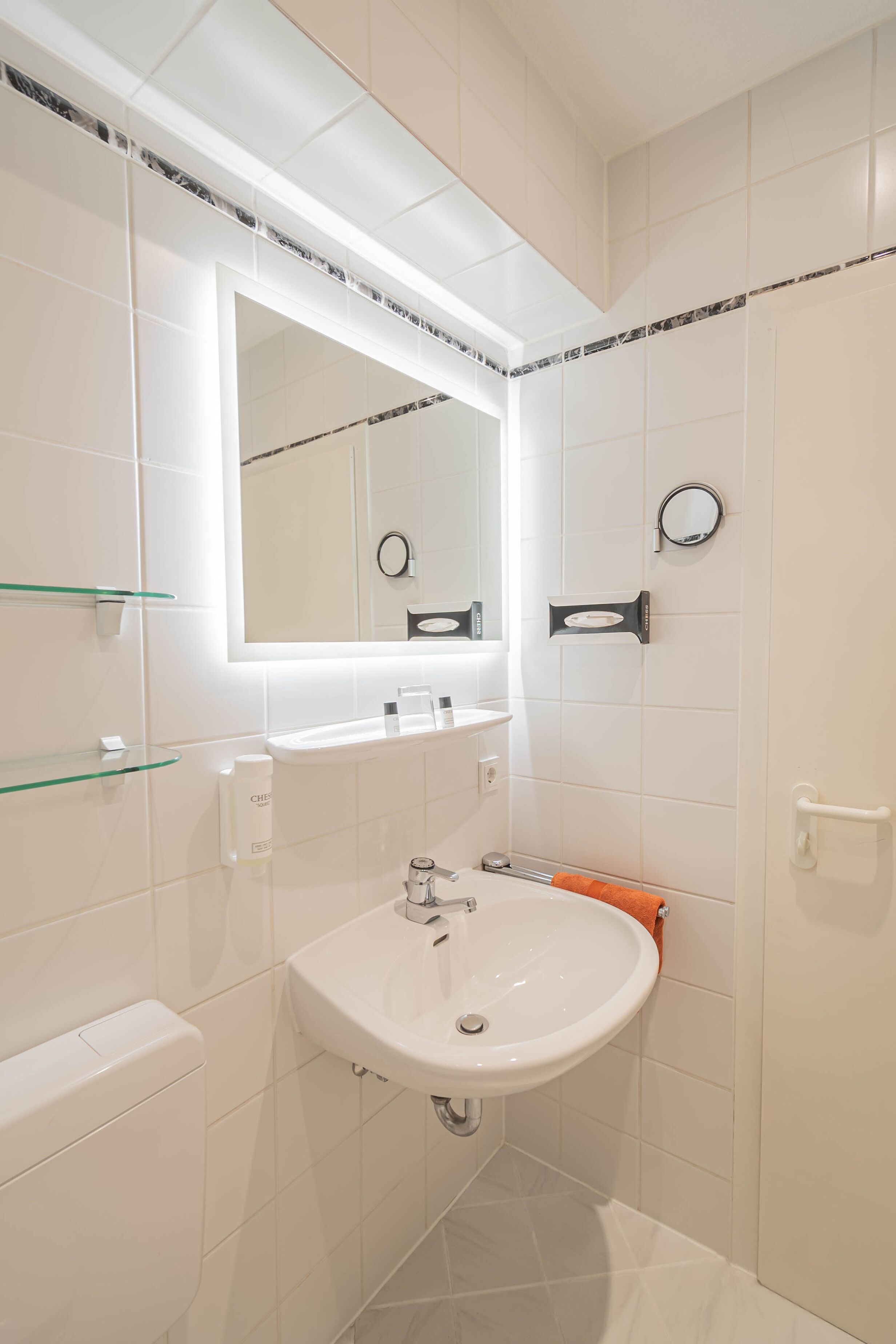 Un baño moderno con lavamanos blanco, espejo y estantes de vidrio.