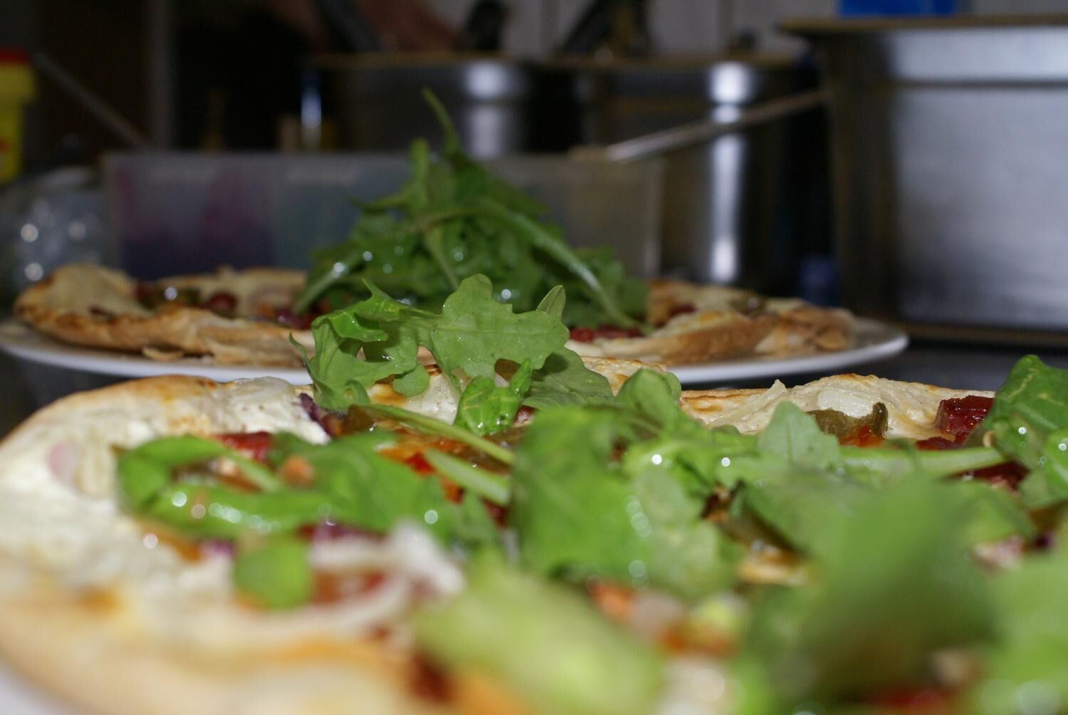 A pizza topped with greens and cheese, with a blurred background of kitchen equipment.