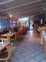 Restaurante com mesas e cadeiras de madeira, piso de azulejos e vista para o exterior através de grandes janelas.