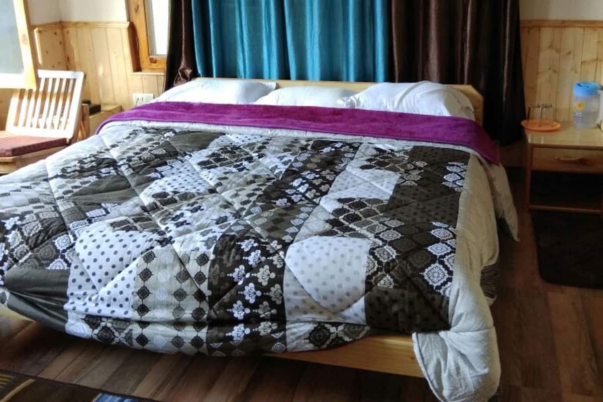 A bed with a patchwork quilt, a wooden chair, a desk with a lamp, and curtains with a leaf pattern.